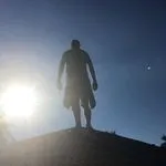 Silhouette of a person standing on a hill or rooftop against a bright blue sky, with the sun shining brightly to the left.