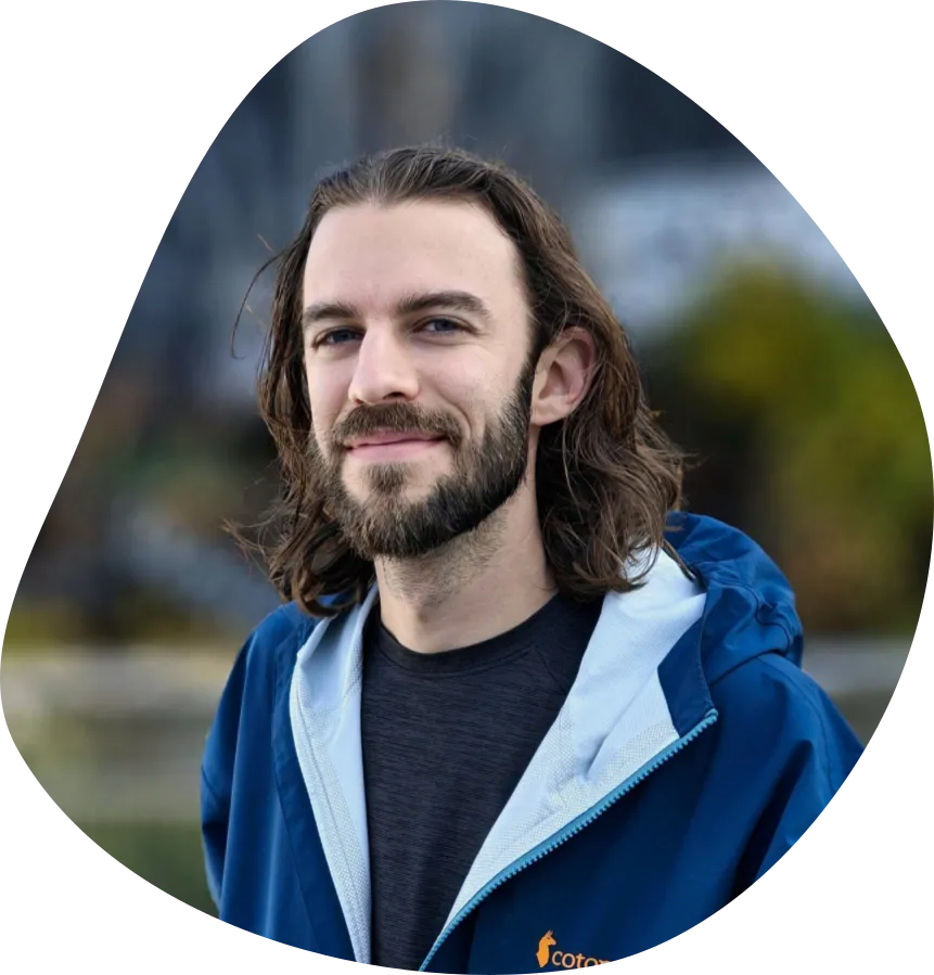 Photograph of Bennett smiling in front of a blurry outdoor background, wearing a blue jacket.
