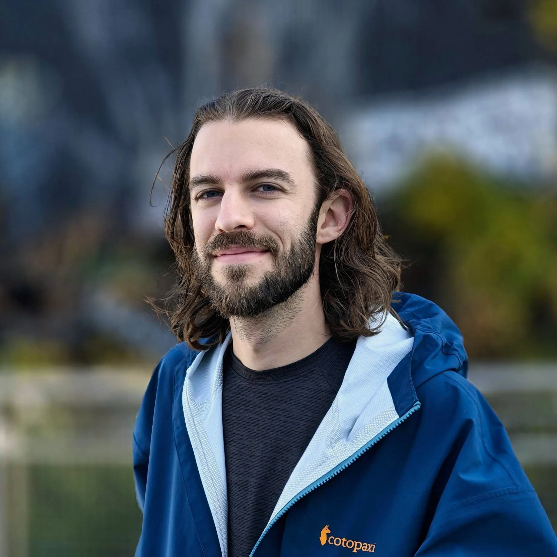 Photograph of Bennett smiling in front of a blurry outdoor background, wearing a blue jacket.
